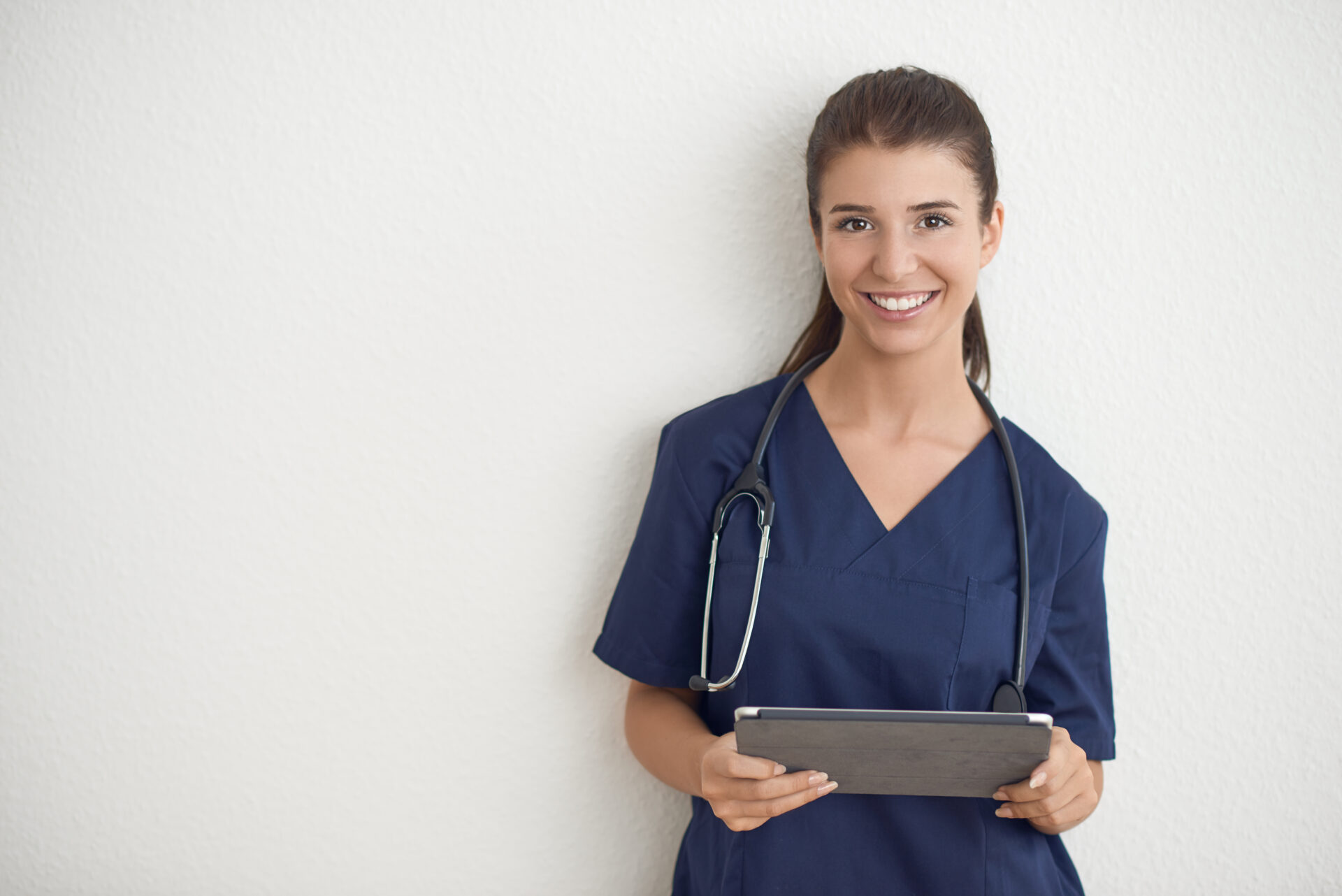 Young woman doctor or nurse in blue scrubs holding a tablet pc standing in front of a white wall